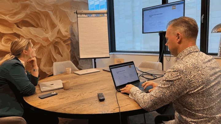 Twee collega's aan een vergadertafel kijken naar de website op een groot scherm en een laptop