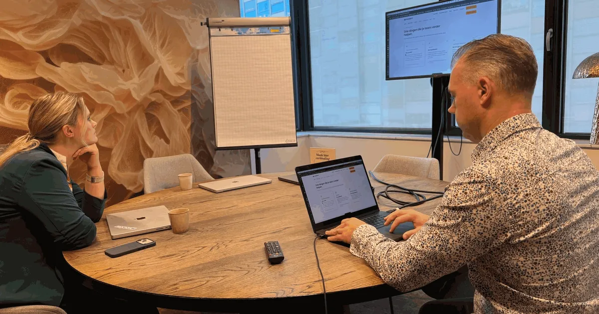 Twee collega's aan een vergadertafel kijken naar de website op een groot scherm en een laptop